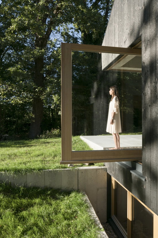 Grande fenêtre pivotante vitrée dans une façade en bois sombre, avec une femme sur le balcon derrière, projet Black House par Buero Wagner, avec charnière pivotante FritsJurgens System M+